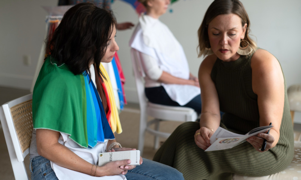 Two individuals seated indoors during a colour analysis session, with one person holding a fan of colour swatches draped across their shoulders and another holding a booklet. Additional colour drapes are visible in the background.